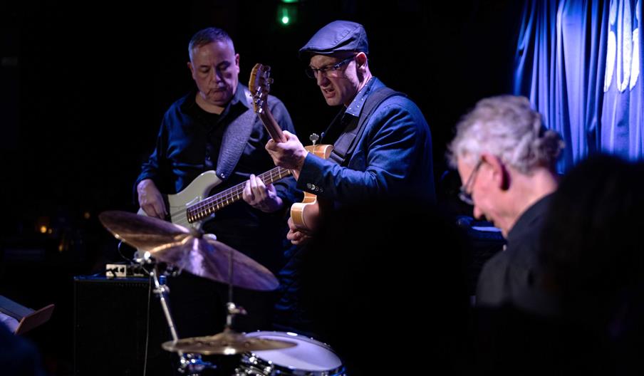 Three musicians perform on stage in a dimly lit jazz club, with one playing electric bass, another playing guitar, and a third on drums in the foregro