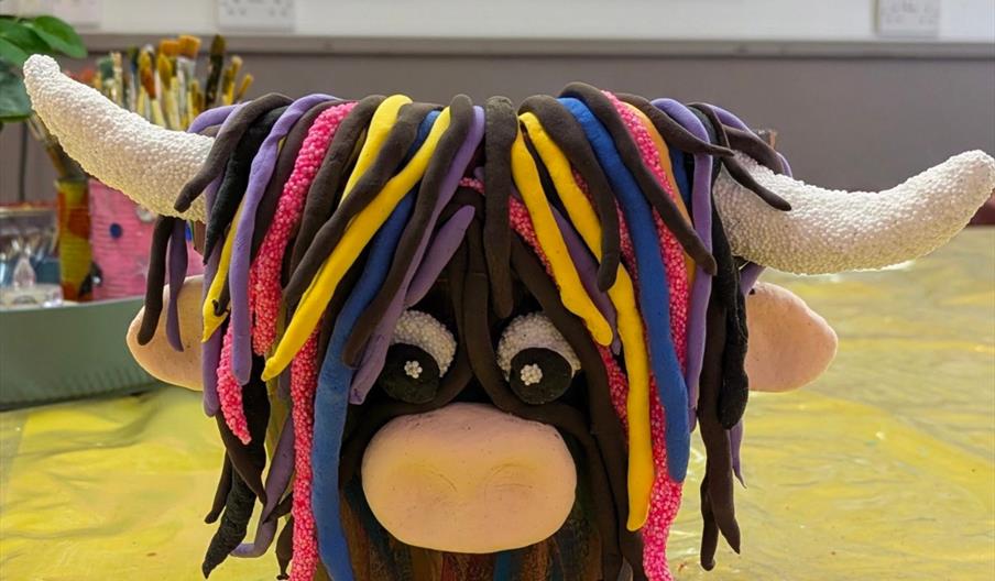 A colourful, handmade clay sculpture of a Highland cow with large white horns and multi-coloured strands for hair, sitting on a yellow-covered table i
