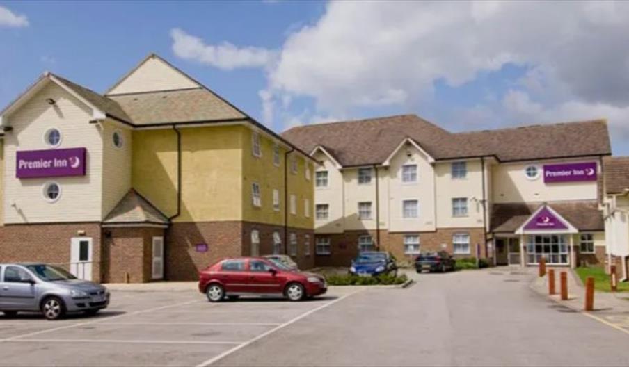 A Premier Inn hotel building with a parking lot in front, several parked cars, and clear signage displaying the hotel’s name on the exterior. The sky