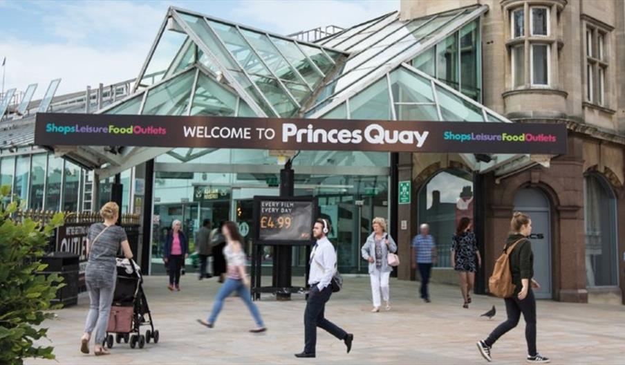 People walk in front of the entrance to Princes Quay shopping center in Hull, which features a glass canopy and a large sign welcoming visitors. Sever