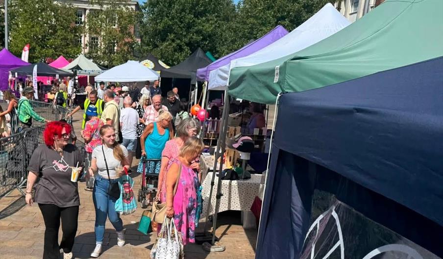 An outdoor market in Queen Victoria Square with green and white tents is set up in front of a large, ornate gothic-style church, with people browsing