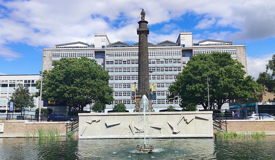 A tall column monument with a statue on top stands in front of a modern office building, flanked by two large trees. In the foreground, there is a pon