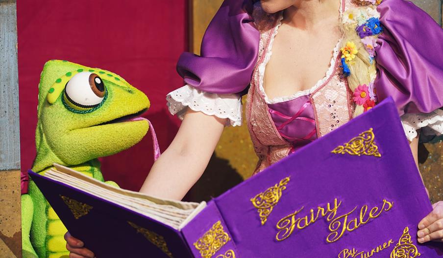 A green chameleon and a girl with a purple dress and long blonde hair look at a fairy tails book