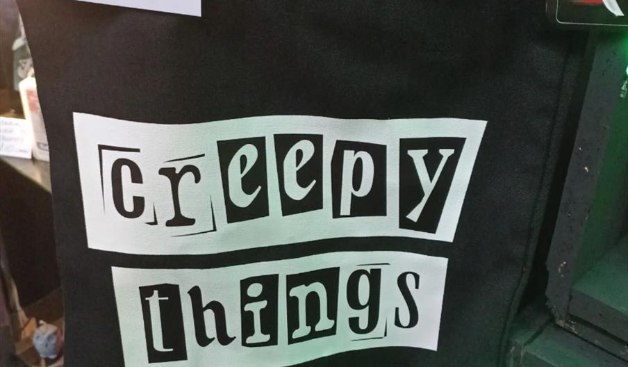 Interior of ReVelation 13. A black tote bag with the words "creepy things" printed on it in a ransom-note style font. A sign above reads "Tote Bags £6