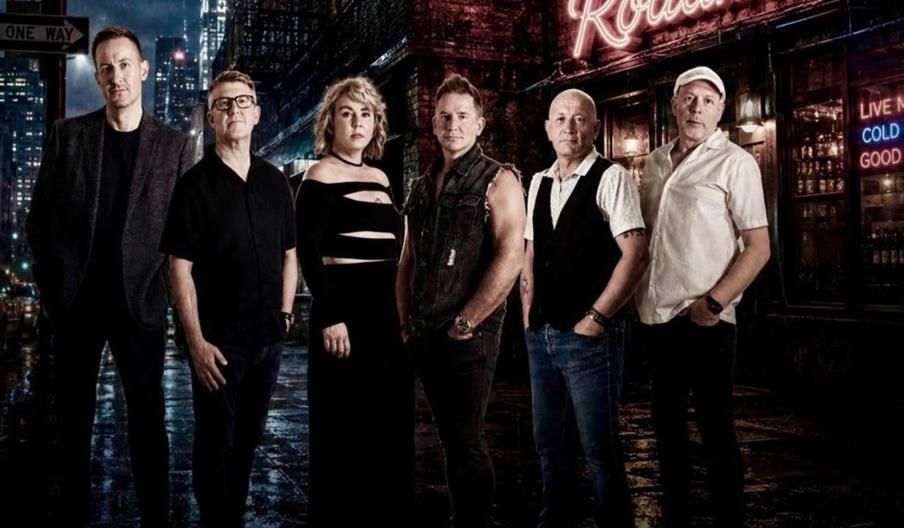 Six people, four men and two women, stand in a row on a wet city street at night under a neon "Roadhouse" sign, with a dark, urban background and a "W