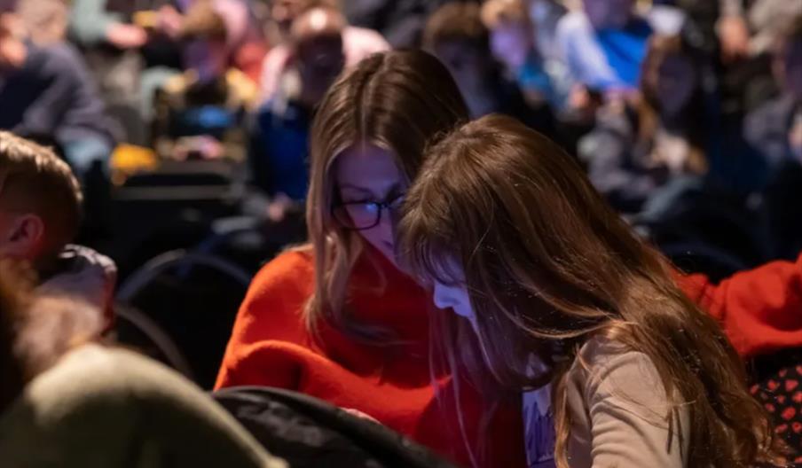 A woman and a young girl sit closely together in an audience, looking down at something in front of them, with many people blurred in the background.