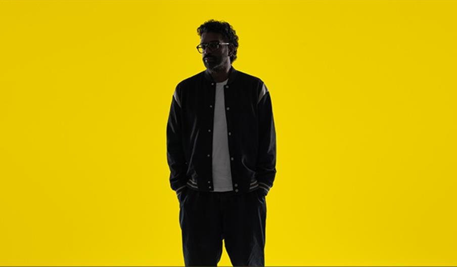 Romesh Ranganathan in a dark jacket, and light shirt stands with his hands in his pockets against a solid bright yellow background.