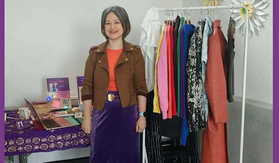 A woman with shoulder-length gray hair stands indoors, smiling, beside a rack of colorful clothes. To her left is a table with a purple patterned clot