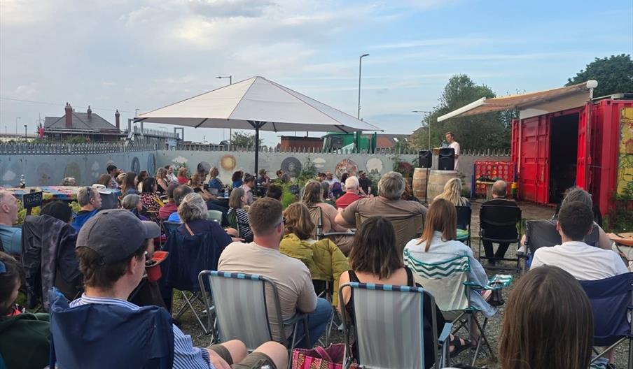 A group of people sit on folding chairs outdoors, facing a person speaking on a small stage set up in front of an open red shipping container, with a