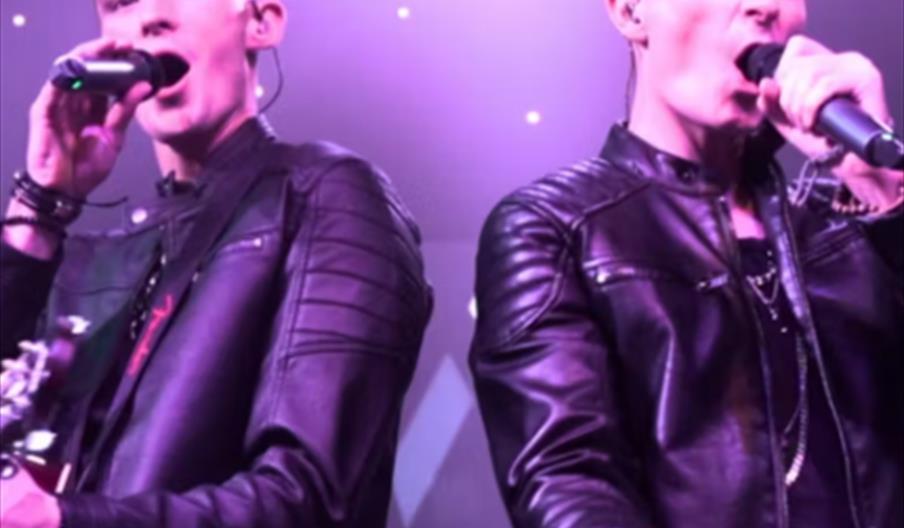 Two male performers in black leather jackets sing into microphones on stage under purple lights, with the bold yellow text "ROX VOX" across the bottom
