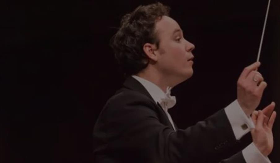 A conductor in a formal black suit and white bow tie leads an orchestra, holding a baton and gesturing with his hands.
