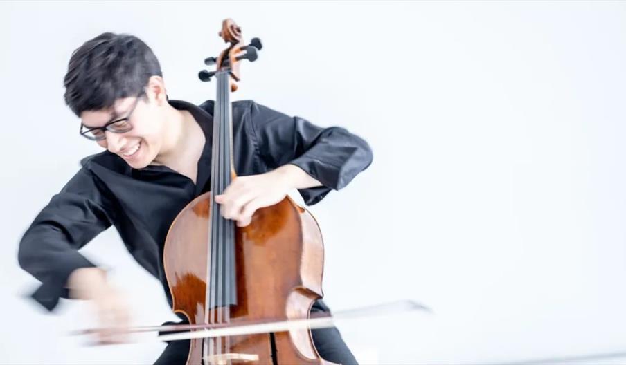 A young person wearing glasses and a black shirt is playing a cello energetically, smiling and captured in motion against a plain light background.