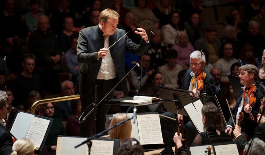 A conductor leads an orchestra during a live performance, with musicians playing string instruments and sheet music visible in front of them; an audie