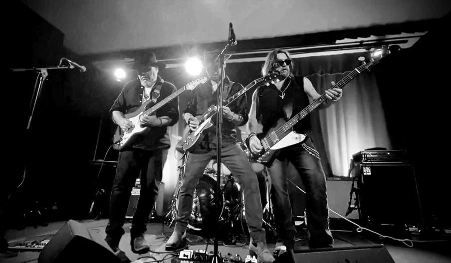 Black and white photo of three musicians playing electric guitars on stage, with a drum set in the background and stage lights shining behind them.