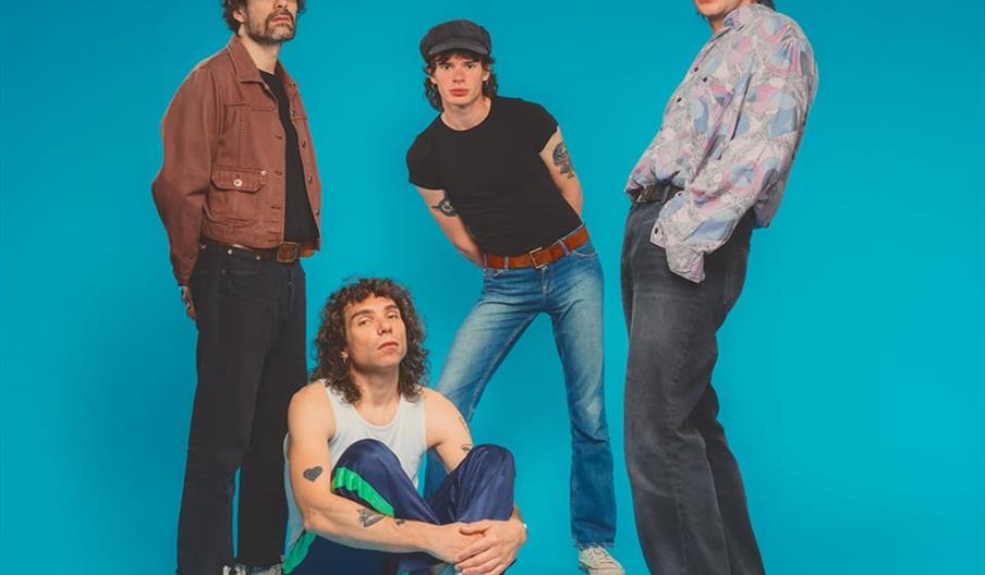 Four men pose against a bright blue background; three are standing in different poses and one is sitting on the floor with his arms around his knees.