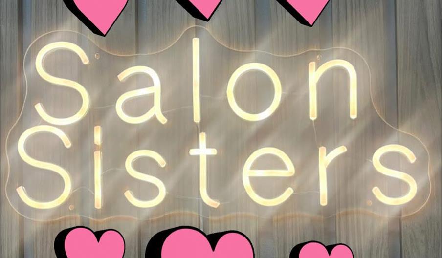 A neon sign on wood paneling displays the words "Salon Sisters" in glowing white letters, surrounded by six large pink hearts with black shadows.