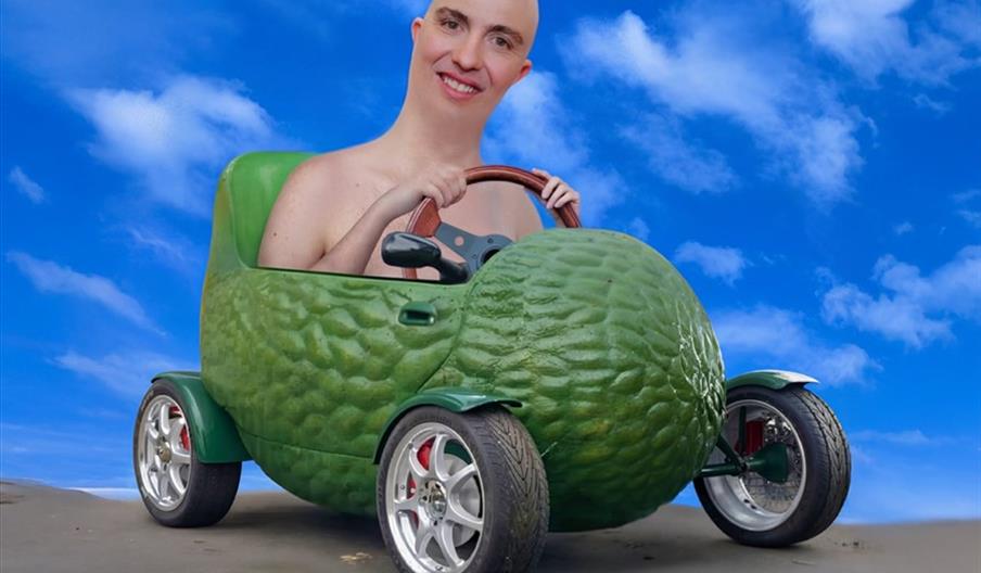 A person with a large smiling head is seated in a small, cartoonish car shaped like a green avocado, set against a bright blue sky with scattered clou