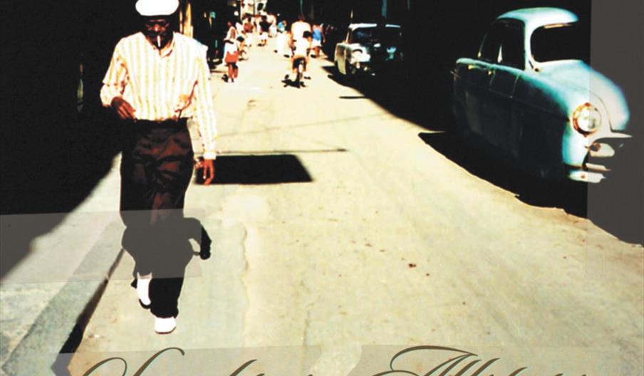 A sepia-toned image of a man wearing a light shirt and white cap walking down a sunlit street with old buildings and a classic blue car; text below ad
