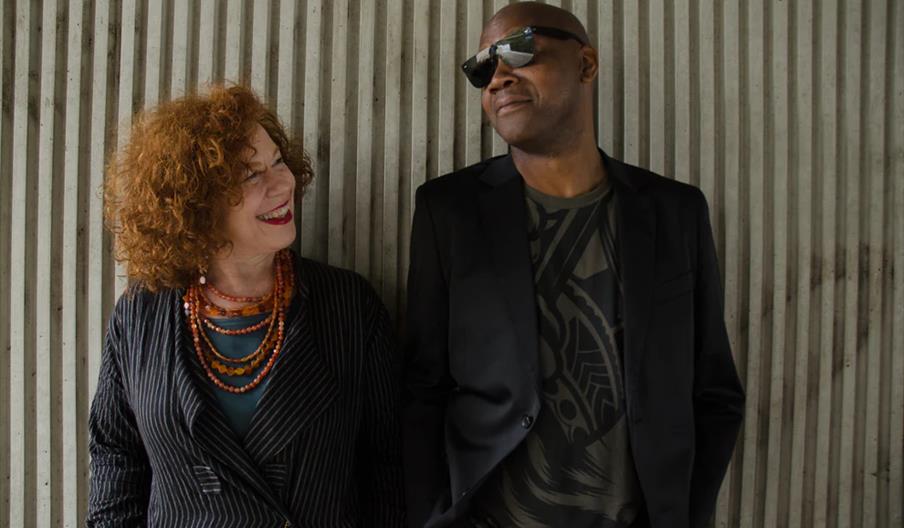 A woman with curly red hair and layered necklaces stands next to a man wearing sunglasses and a black blazer, both smiling at each other in front of a