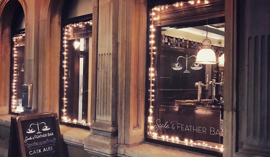 Street view of the Scale & Feather Bar at night, showing its arched windows outlined with string lights, warm interior lighting, and a chalkboard sign
