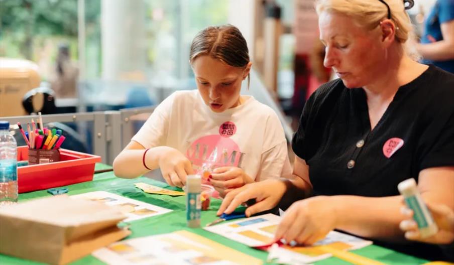 A woman and a young girl sit at a table covered with craft supplies, focusing on assembling or decorating paper projects. The background shows a brigh
