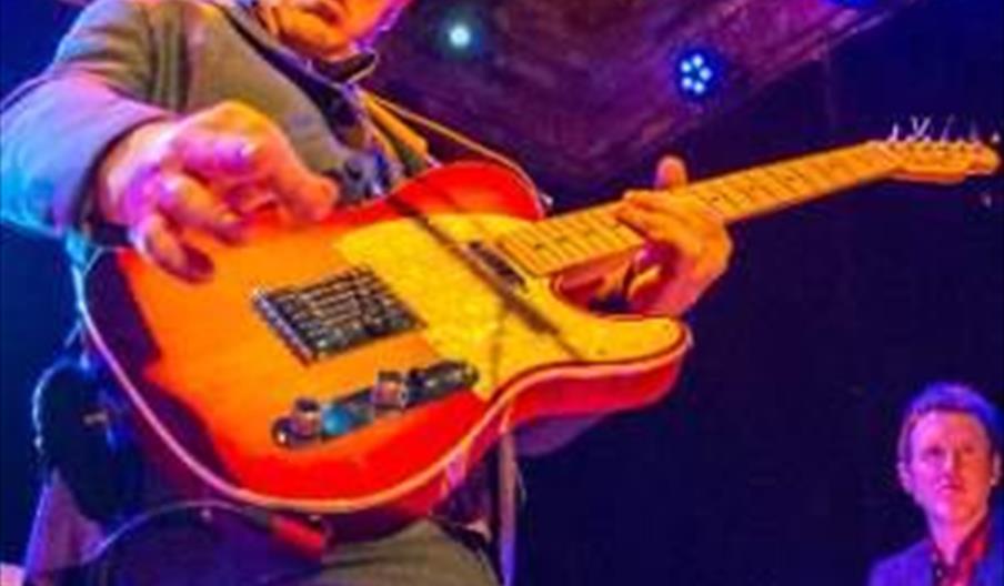 A musician in a blue suit passionately plays an electric guitar on stage, while another person plays a keyboard in the background under colorful stage
