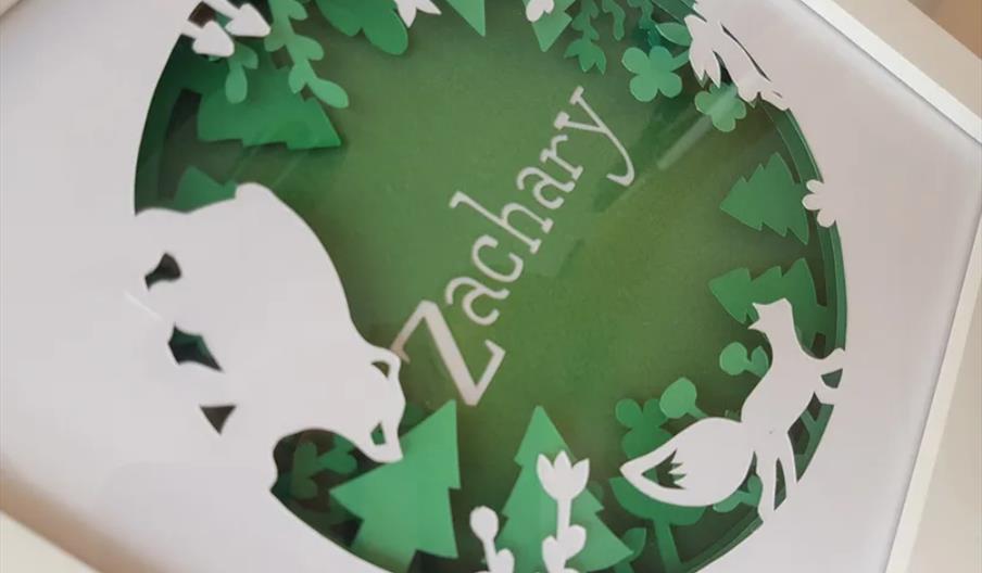 A framed paper art piece featuring the name "Zachary" in the center, surrounded by layered green trees and white animal silhouettes, including a bear