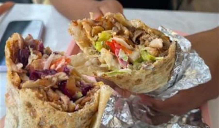 Two people holding halves of a loaded wrap or burrito filled with colorful vegetables, meat, and other ingredients, partially wrapped in foil over a t