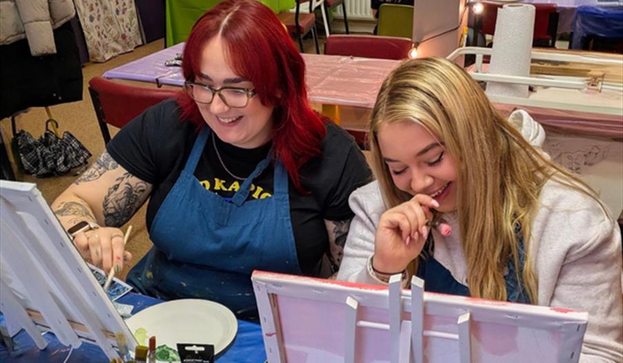 Two women sit side by side at a table, smiling and laughing while painting on canvases with paintbrushes, surrounded by art supplies in a creative stu