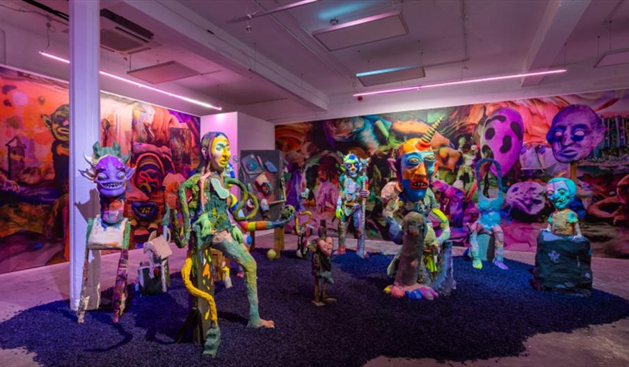A brightly lit art gallery within Humber Street Gallery featuring colourful, surreal humanoid sculptures arranged in a circle on a bed of dark gravel,