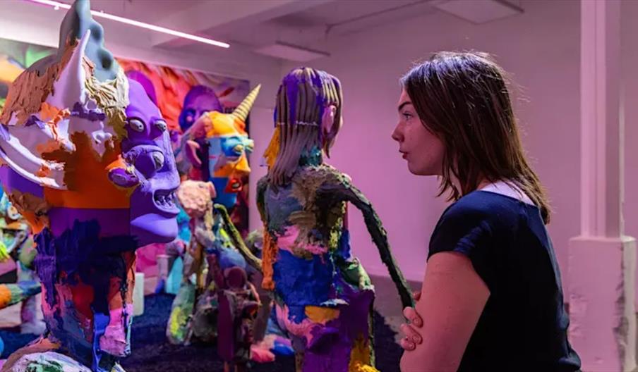 A woman stands in Humber Street Gallery, closely examining a colorful, abstract sculpture among several other vibrant, eclectic humanoid figures. The