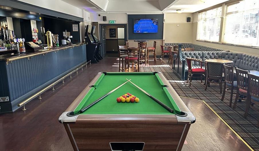 A pool table with neatly racked balls and two cues crossed on top is positioned in the foreground of a spacious pub, featuring a long bar on the left,