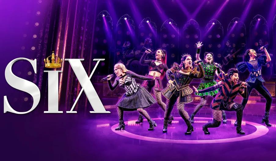 Six women in colorful, stylized costumes perform energetically on stage under vibrant purple lighting, with the large text "SIX" and a gold crown grap