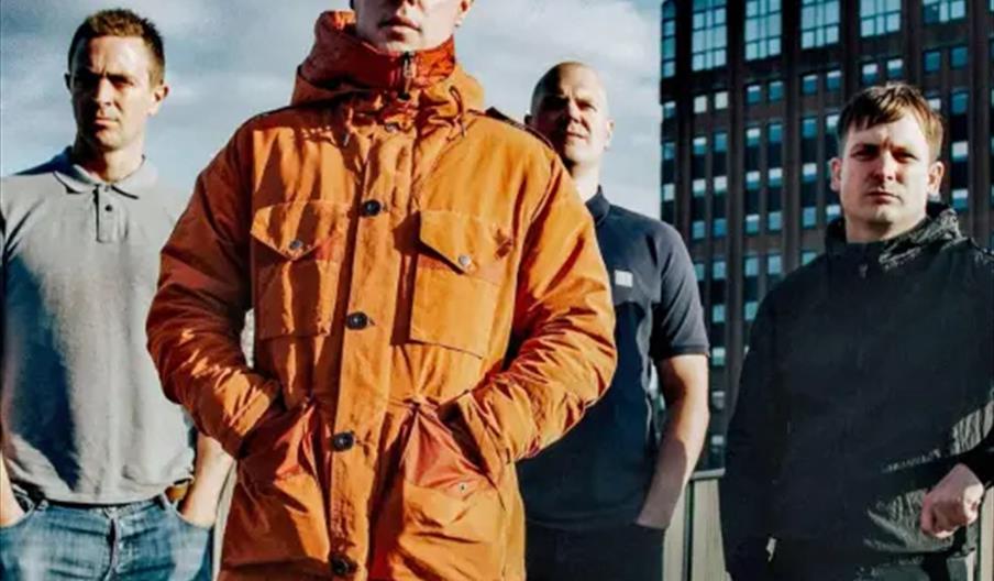 Four men stand outdoors on a bridge, with one man in an orange jacket in the foreground and three others behind him; a modern building and a cloudy sk