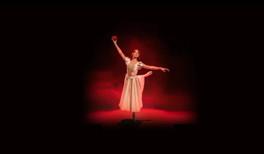 A ballerina in a white dress balances on one foot on stage, holding a red apple aloft with her right hand and extending her left arm gracefully, lit b