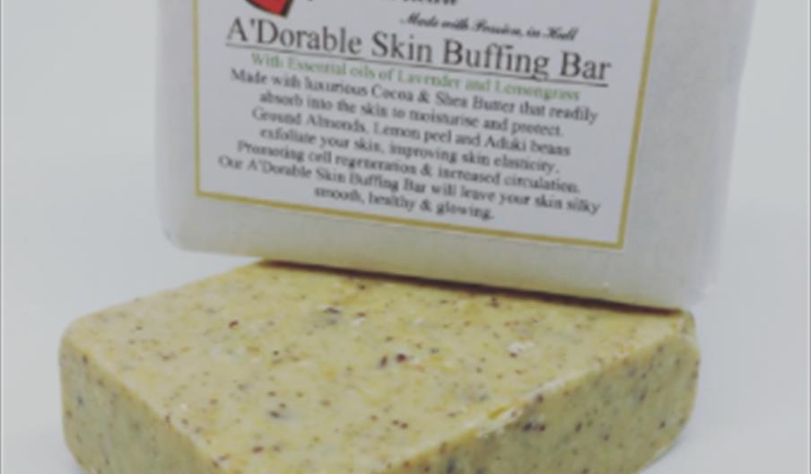 A handmade rectangular skin buffing bar with visible exfoliating particles sits in front of its white paper packaging, which features product informat