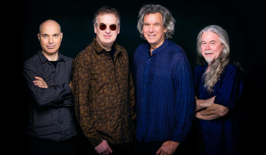 Four men stand side by side against a dark background, all smiling; one is bald, two have short hair, and one has long gray hair and a beard.