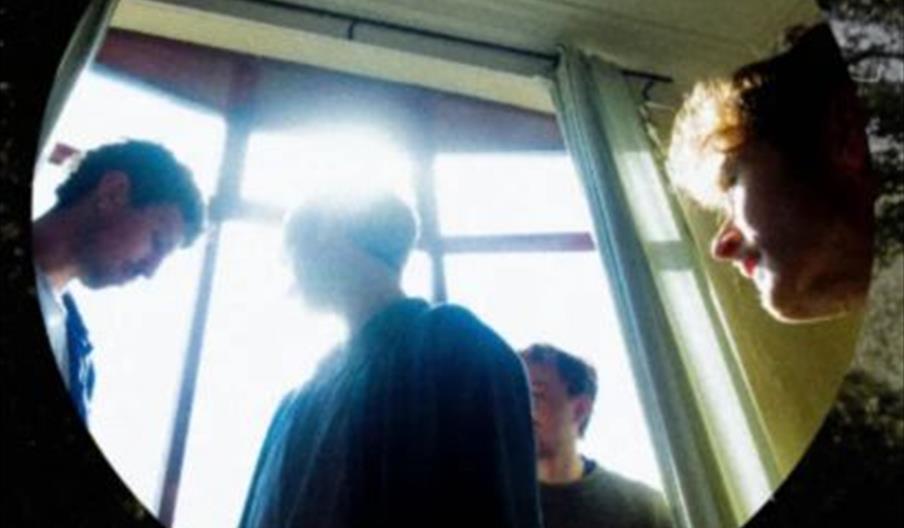 Four people stand in a sunlit room near a large window, with bright light streaming in behind them. The image is framed in a circular shape, and the w