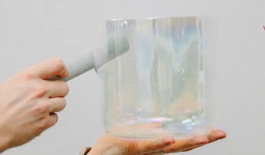 A person holds a clear, iridescent crystal singing bowl in one hand and rubs the rim with a mallet using the other hand.