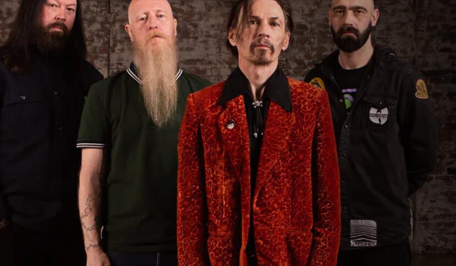 The group Space, four men stand indoors in front of a brick wall; the man in front wears a bright red patterned jacket, while the others wear darker,