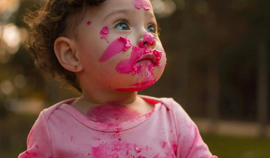 A young child wearing a pink shirt has bright pink cake or frosting smeared all over their face and neck, looking upward with a thoughtful expression.