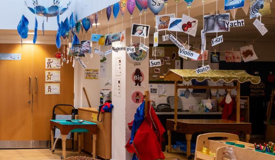A colorful classroom decorated with alphabet letters, pictures, and words hanging from the ceiling; children's artwork and learning materials are disp