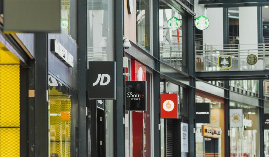 Modern indoor shopping centre corridor with branded store signs including JD, Boux Avenue, Vodafone and Starbucks, featuring glass walkways, retail un
