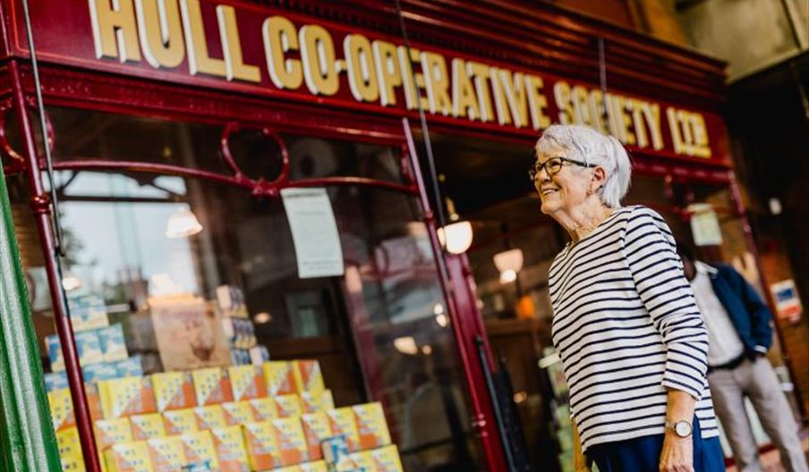 An older woman with short gray hair and glasses, wearing a striped shirt, stands smiling outside a shop with a red sign reading "Hull Co-Operative Soc