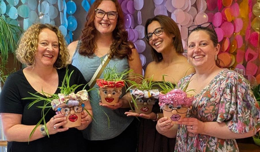 Four women smile and pose together indoors, each holding a decorated plant pot shaped like a face with googly eyes, lips, and fabric accessories as ha