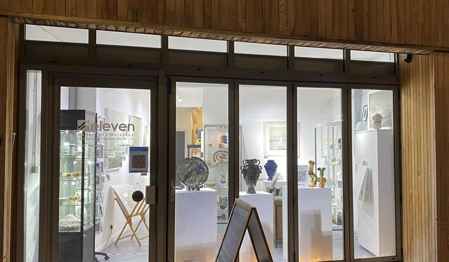 A well-lit front of Studio Eleven with large glass windows and a wooden exterior, displaying ceramic and glass art pieces on pedestals inside, and a s