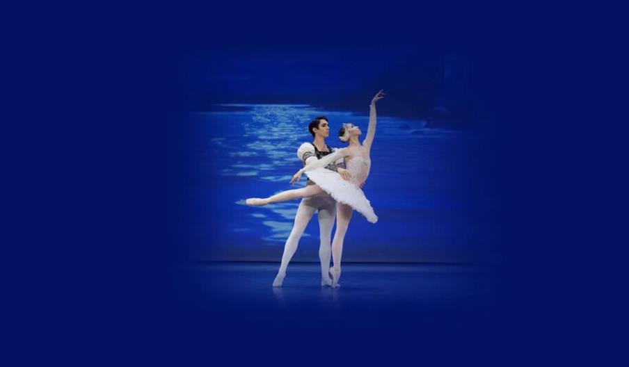 Two ballet dancers perform an elegant pose on stage, with the man supporting the woman as she extends one leg and one arm gracefully. The background s