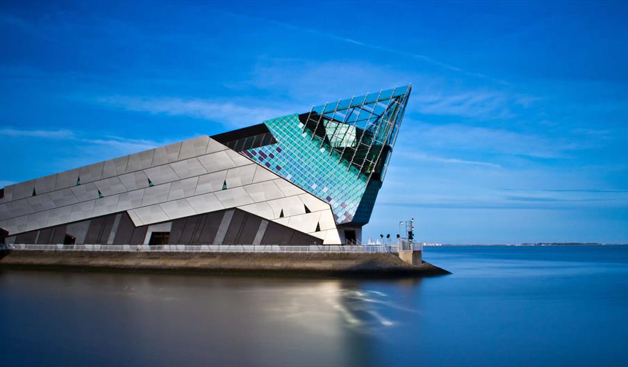 An angular, modern building with metallic and glass surfaces sits on the edge of calm water under a clear blue sky. The Deep features sharp, geometric