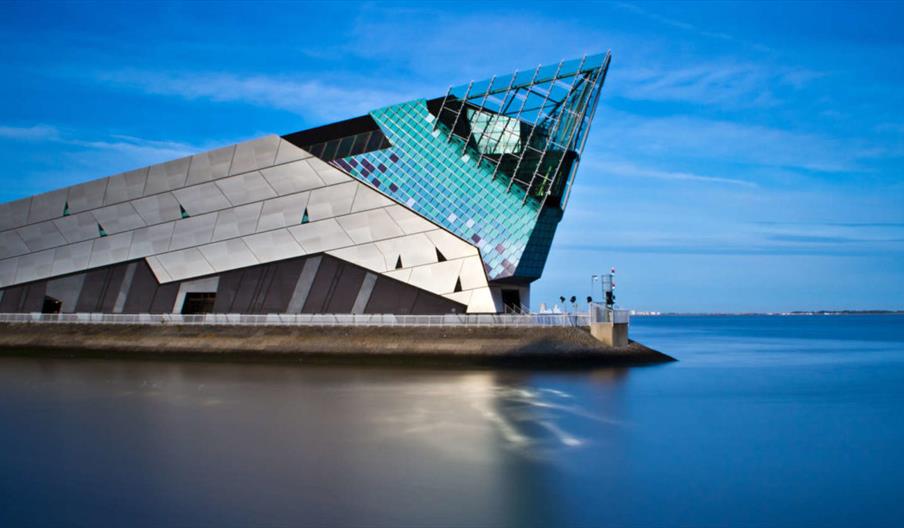 Exterior of The Deep, a modern, angular building with metallic and glass panels sits at the edge of calm blue water under a clear sky, featuring an ir