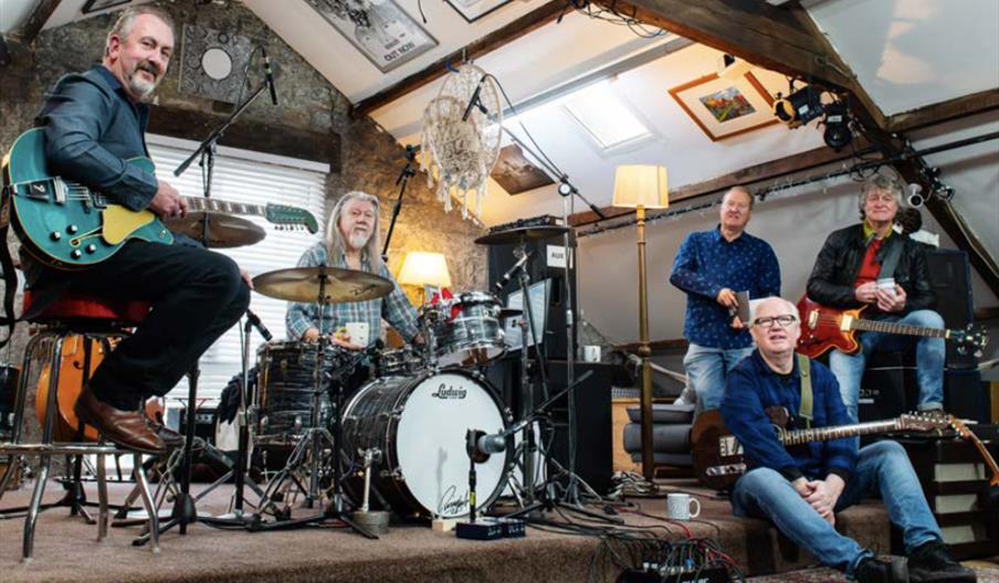 Five middle-aged men are gathered in a cozy, attic-like room, surrounded by musical instruments and equipment. Three are seated with guitars, one sits